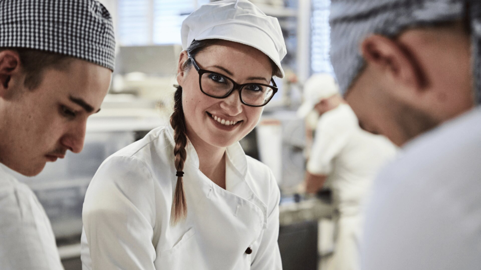 Eine lächelnde Frau mit Brille und weißer Kochuniform steht in einer Küche mit zwei anderen Köchen, die sich auf ihre Arbeit konzentrieren. Der Hintergrund zeigt eine geschäftige, gut beleuchtete professionelle Küchenumgebung.