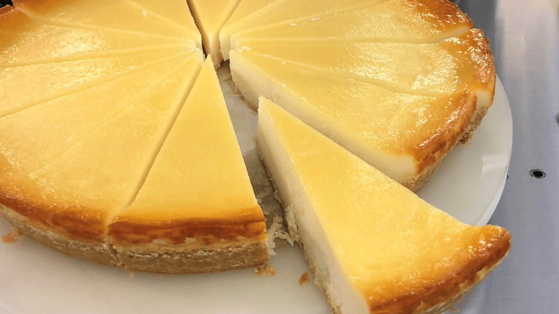 A creamy cheesecake cut into several slices on a white plate, with one slice slightly pulled out from the rest. The cheesecake has a golden-brown edge and a smooth, glossy surface.