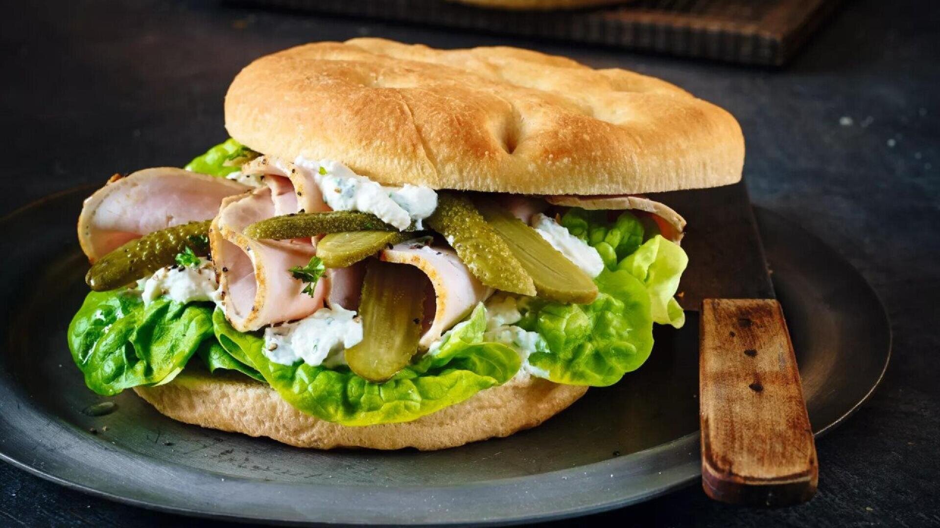 A sandwich on a round bread roll filled with leafy lettuce, sliced deli meat, creamy white spread, and sliced pickles, served on a metal plate with a wooden-handled knife.