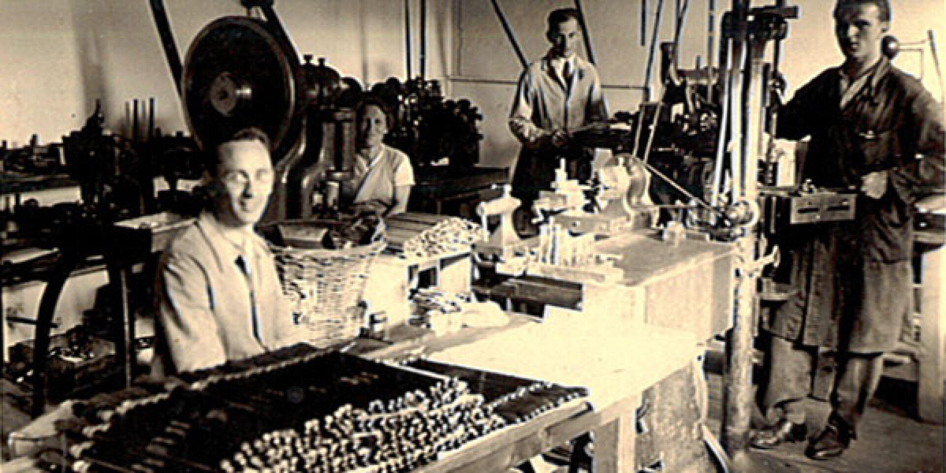 Vier Personen arbeiten in einer alten Fabrikumgebung, umgeben von Maschinen und Werkzeugen. Eine Person sitzt an einem Schreibtisch im Vordergrund mit montierten Teilen, während andere im Hintergrund stehen und Geräte bedienen.