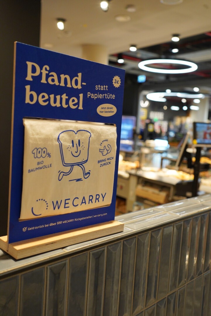 A reusable cotton bread bag with a smiling bread illustration is displayed on a stand in a bakery, promoting its use instead of paper bags. The sign is in German and highlights environmental benefits.