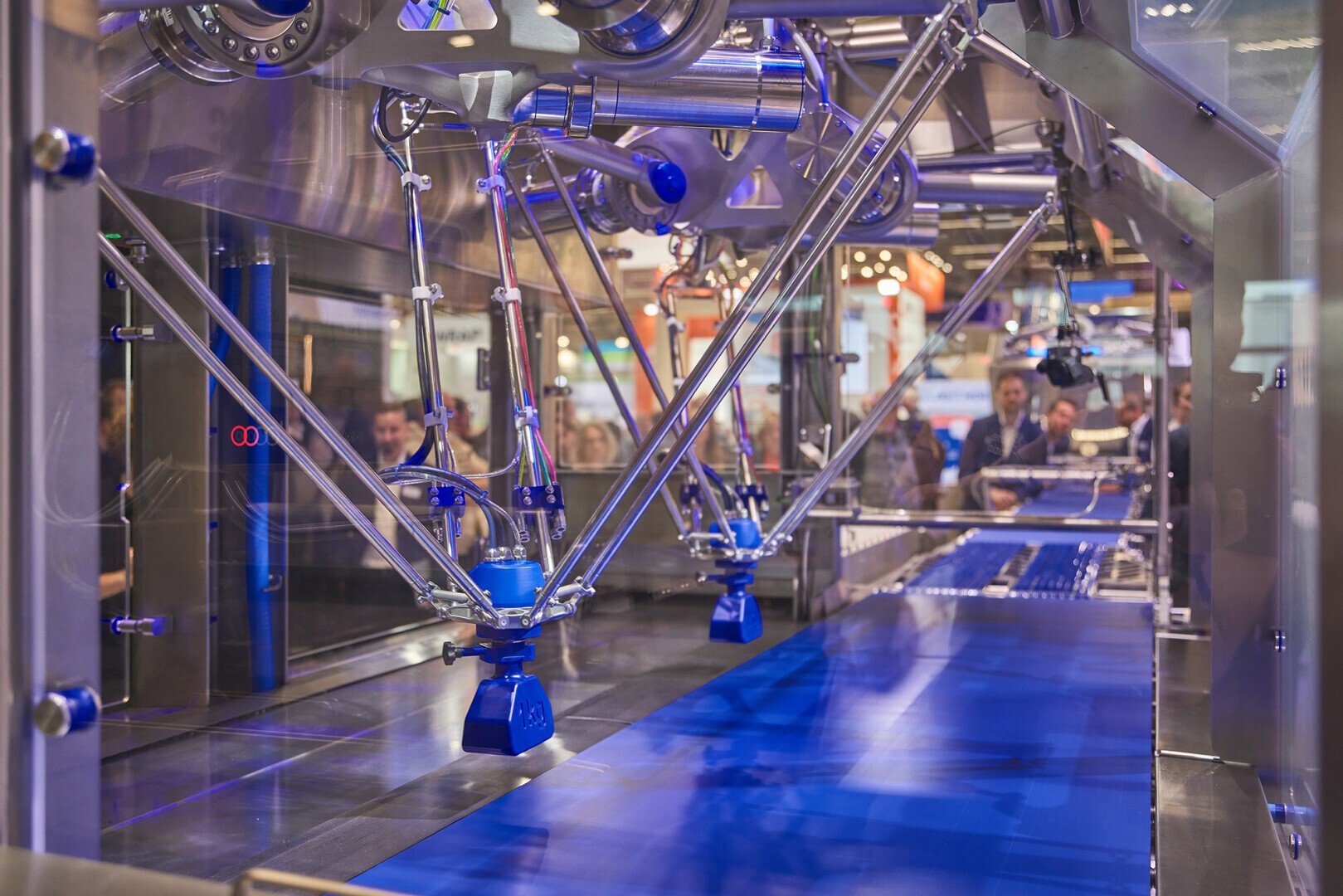 Ein Roboterarmsystem mit mehreren blauen und silbernen Armen arbeitet an einem blauen Förderband in einer Fabrikumgebung, während Menschen durch eine Glasplatte im Hintergrund zuschauen.