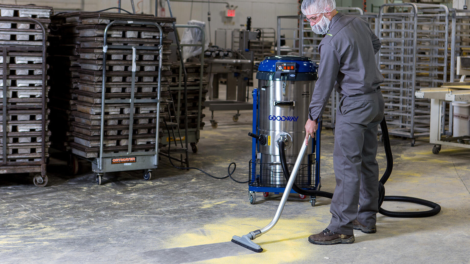 A worker wearing protective clothing and a hairnet uses an industrial vacuum to clean yellow powder from the floor of a facility with metal racks and baking trays in the background.