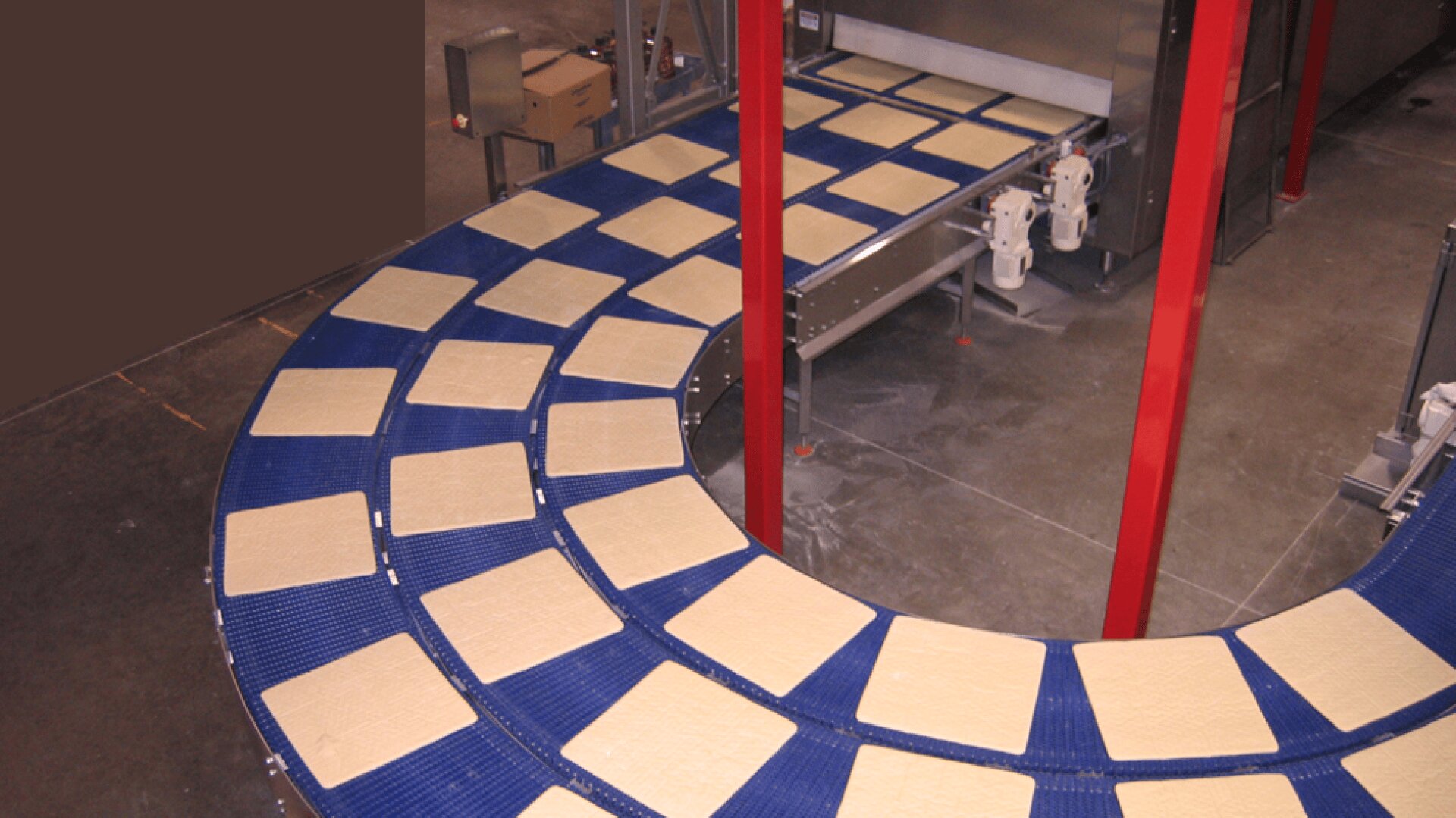 Squares of uncooked pizza dough move along a curved blue conveyor belt in a food processing facility, heading toward a large industrial oven or machine.