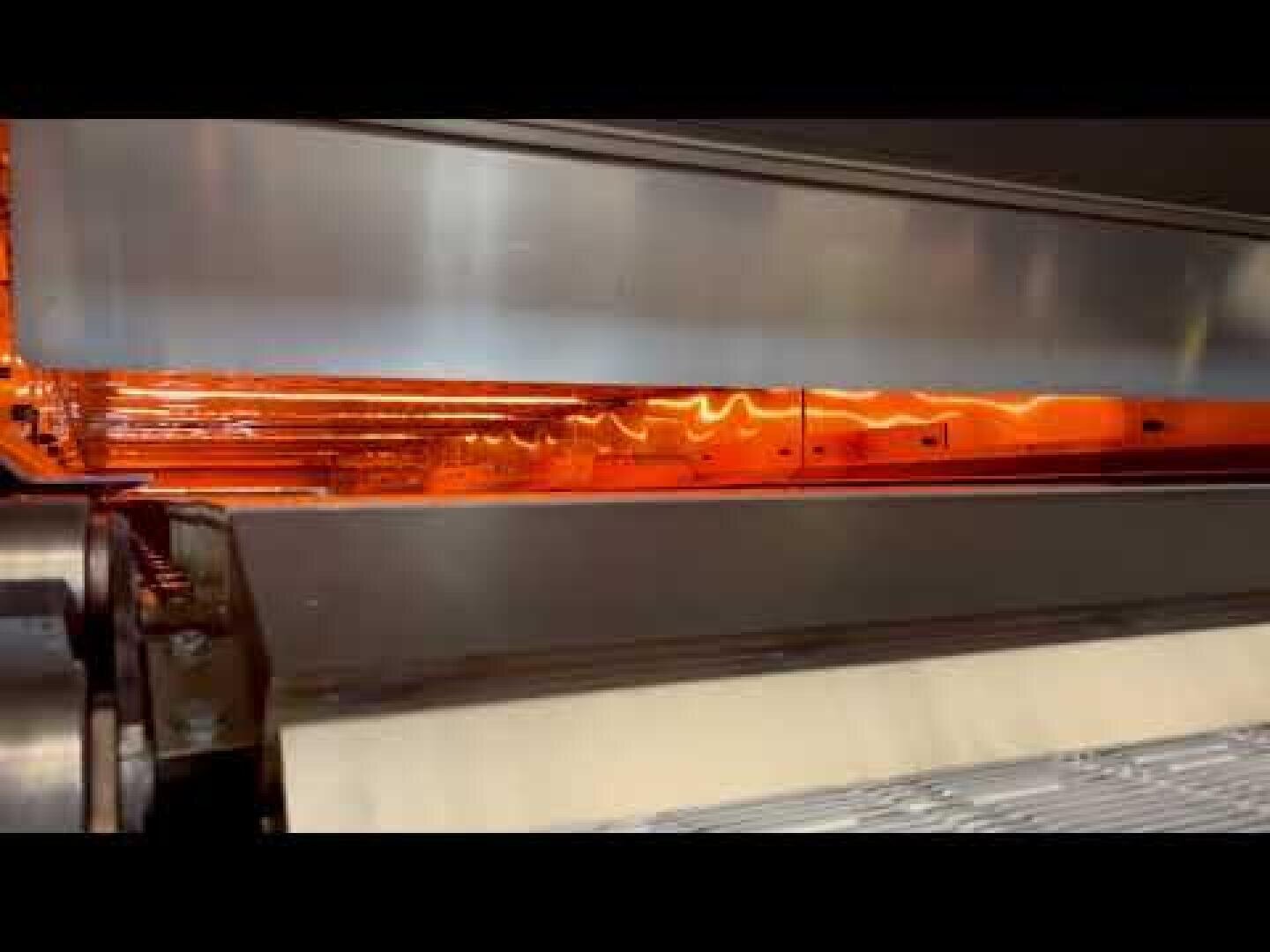 Eine Nahaufnahme einer Industriemaschine mit leuchtend orangefarbenen Heizelementen im Inneren, Metallrollen und einem Förderband, das sich durch die Anlage bewegt.