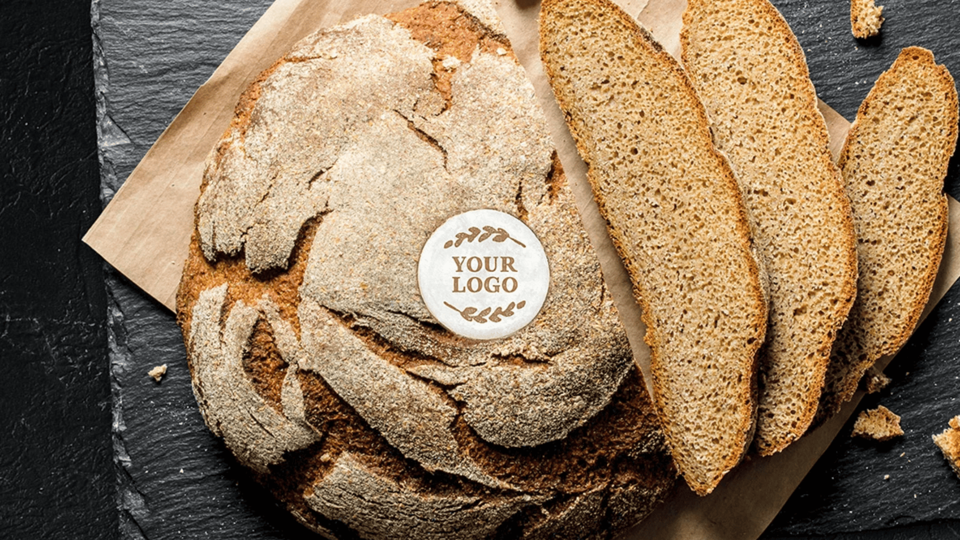 Ein runder, rustikaler Brotlaib mit einem Your Logo-Etikett auf der Oberseite, teilweise aufgeschnitten, mit drei daneben liegenden Scheiben auf Pergamentpapier und einer dunklen Schieferoberfläche.