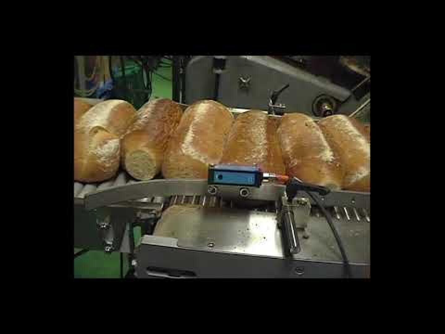 Several loaves of bread covered with flour are moving along a conveyor belt in a factory or industrial setting, with a monitoring device attached to the machinery.