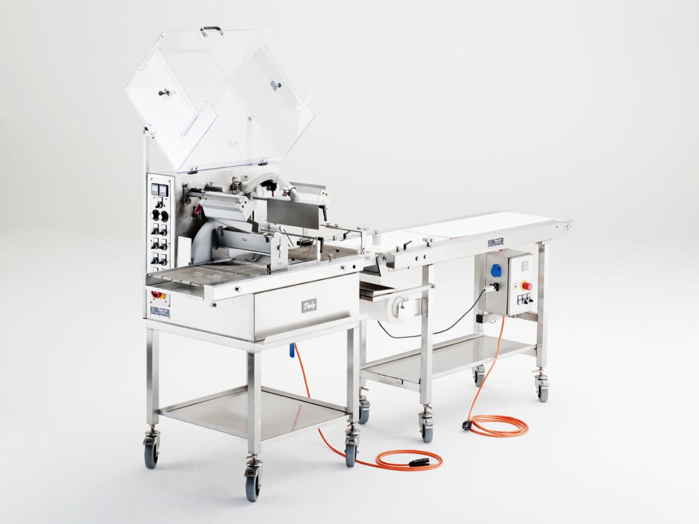 Eine industrielle Lebensmittelverarbeitungsmaschine aus rostfreiem Stahl mit Schalttafeln, einem Förderband und orangefarbenen Stromkabeln auf Rädern, dargestellt in einer sauberen, weißen Studioumgebung.