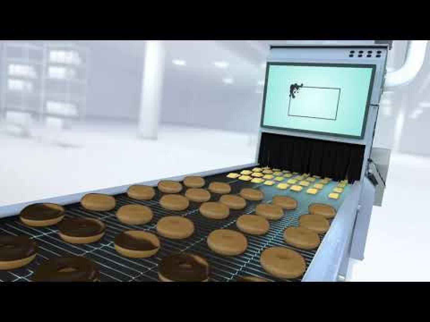 Ein Förderband transportiert Reihen von Donuts unter einer Maschine mit einem Bildschirm, der ein Donut-Bild anzeigt, was auf einen automatisierten Qualitätskontroll- oder Inspektionsprozess in einer Fabrik schließen lässt.