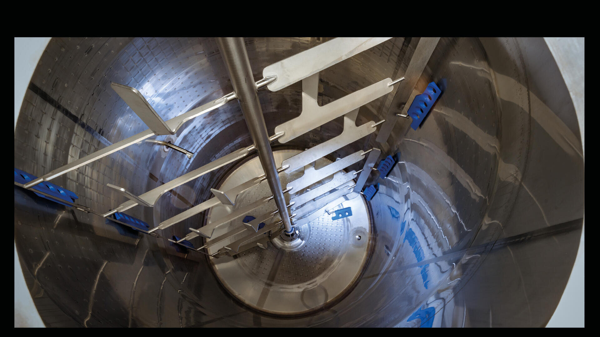 Blick von oben ins Innere eines großen industriellen Mischbehälters aus rostfreiem Stahl. Zu sehen ist eine zentrale vertikale Welle mit angebrachten horizontalen Schaufeln und blauen Kunststoffteilen, die zum Mischen oder Verarbeiten von Materialien verwendet werden.