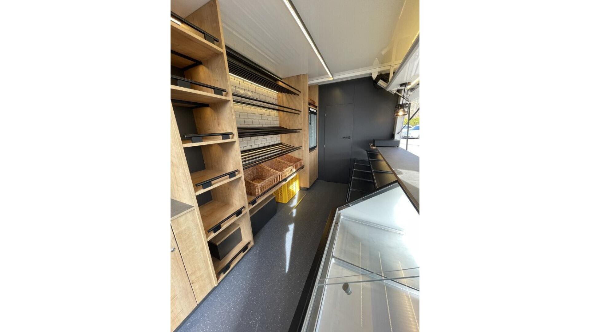 Interior of an empty bakery or deli shop with wooden shelves, bread baskets, a glass display case, and a counter. Sunlight shines in from the open service window on the right.