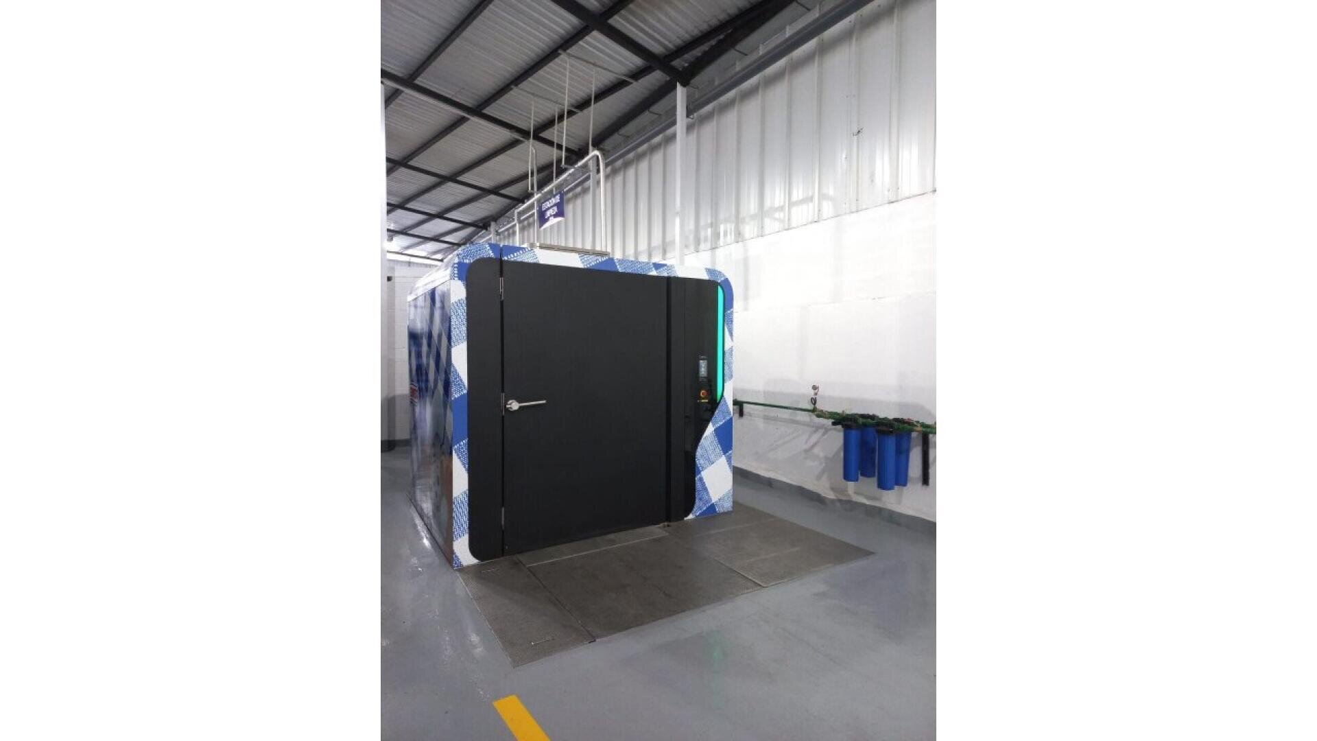 Eine große, schwarze, kastenförmige Industriemaschine mit einem gemusterten Äußeren steht in einer sauberen Halle mit grauem Boden, weißen Wänden und einer hohen Decke. An der Wand daneben sind blaue Filterkanister angebracht.
