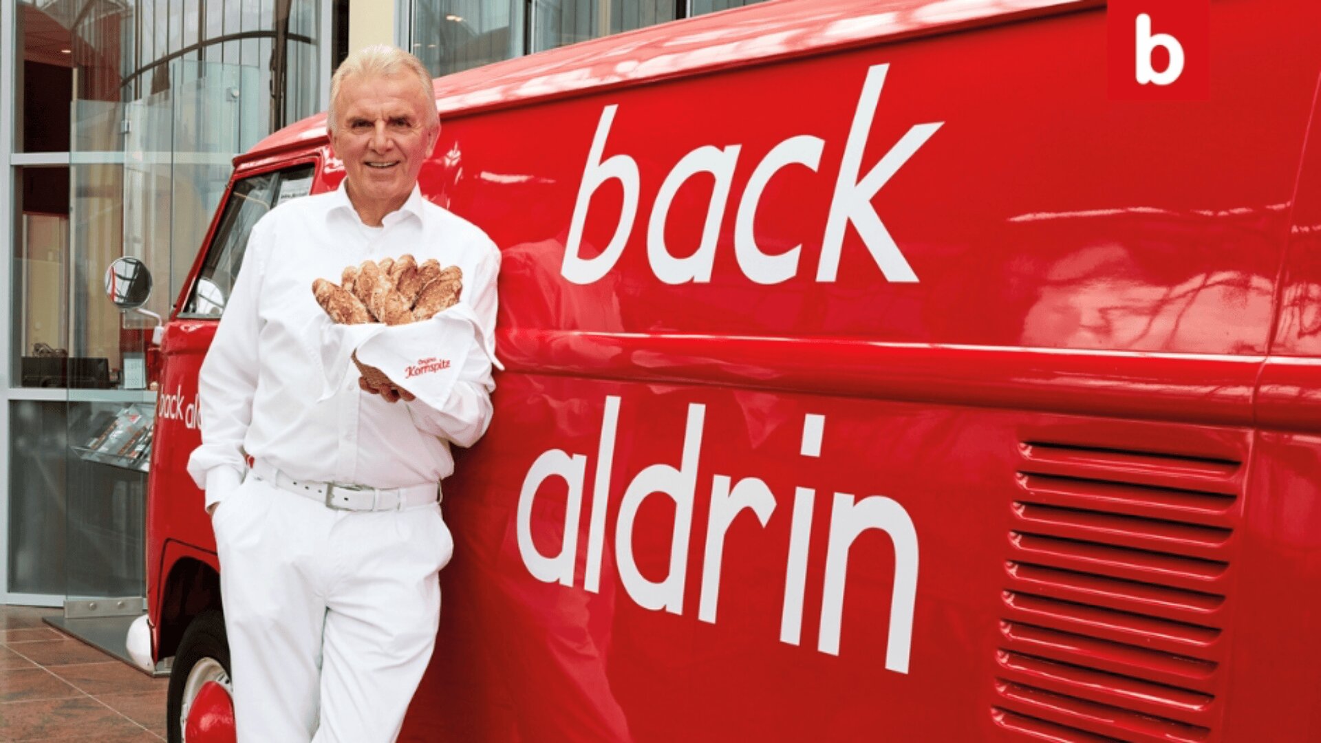Ein älterer Mann in weißer Kleidung hält einen Korb mit Brötchen in der Hand, lächelt und lehnt sich an einen roten Lieferwagen, auf dessen Seite die Aufschrift Back Aldrin steht.