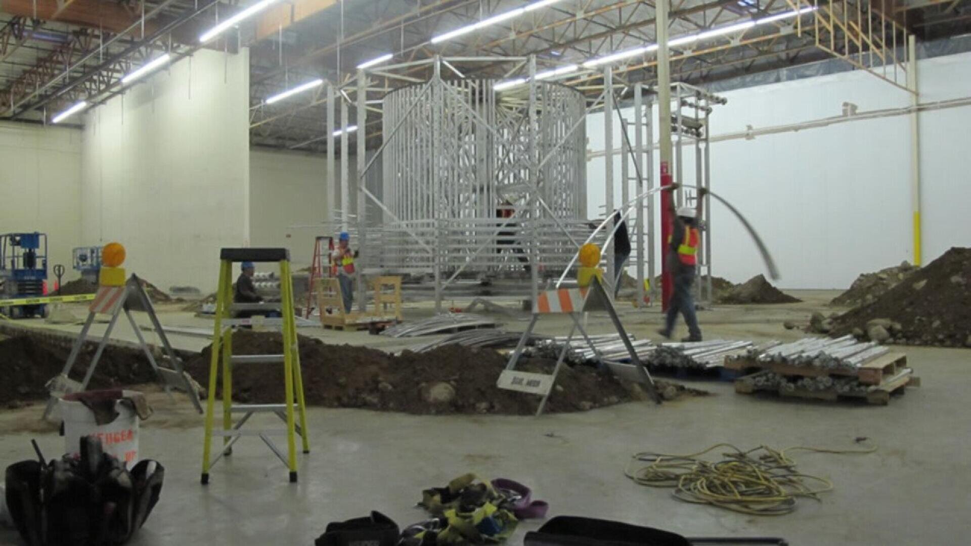 Bauarbeiter in Sicherheitswesten arbeiten in einer großen Lagerhalle, umgeben von Erdhaufen, Metallträgern, Leitern und Baumaschinen. Im Hintergrund steht eine teilweise errichtete Metallrahmenkonstruktion.