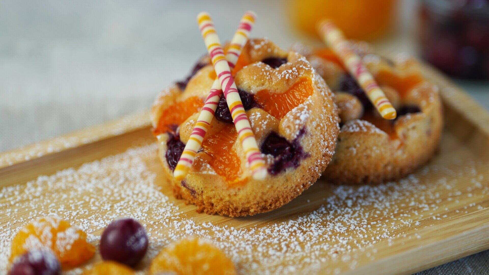 Zwei kleine Obsttörtchen, belegt mit Mandarinenscheiben, Kirschen und dekorativen Schokoladenstiften, bestäubt mit Puderzucker auf einem Holztablett, mit zusätzlichen Früchten und Zucker bestreut.