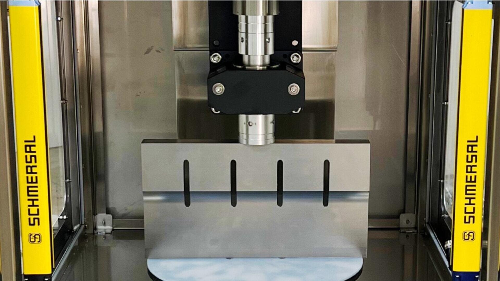 Nahaufnahme einer Industriemaschine mit Sicherheitsschranken mit der Aufschrift Schmersal auf beiden Seiten, die eine Metallpresse oder ein Schneidwerkzeug über einer dicken, gerillten Metallplatte zeigt.