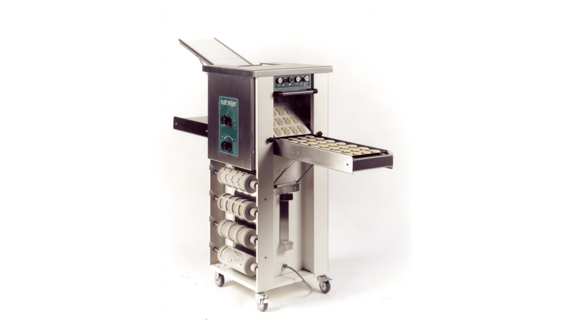 A commercial dough processing machine with rollers and a conveyor belt, dispensing evenly spaced pieces of dough onto a tray. The machine is metallic and stands on wheels for mobility, with various attachments stored underneath.