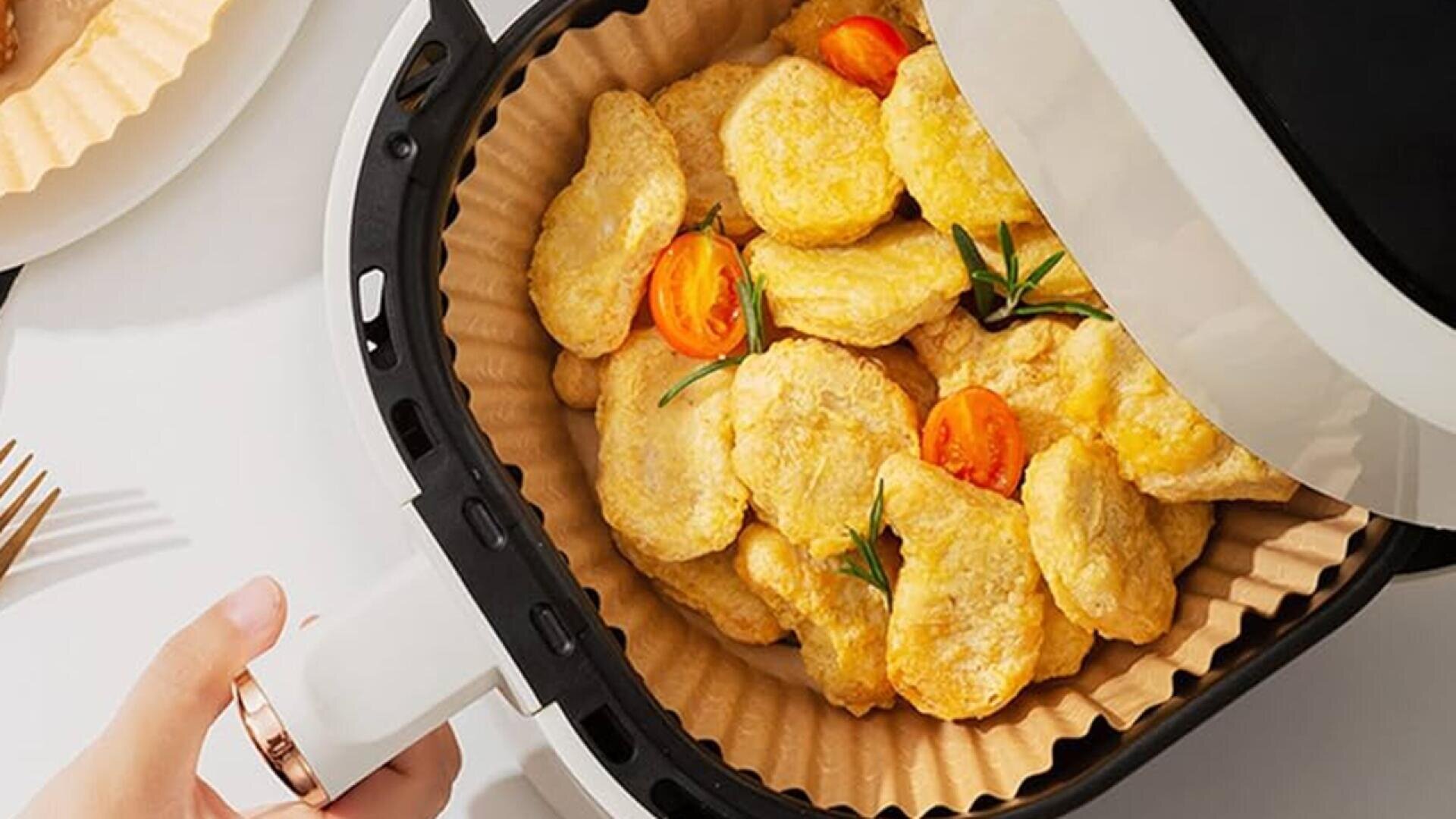 A hand holding an air fryer basket filled with golden chicken nuggets, garnished with sliced cherry tomatoes and rosemary, on a parchment paper liner. Part of a plate and utensil are visible on the white table.