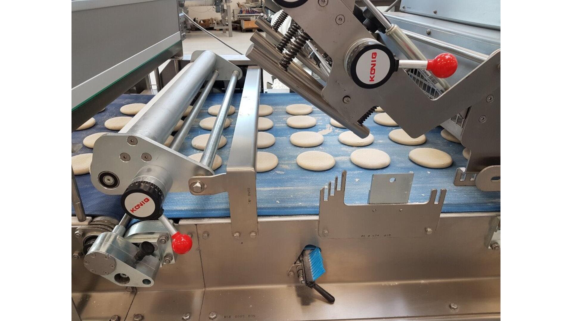 Auf einem blauen Förderband sind Plätzchenteigscheiben aufgereiht, die durch eine industrielle Backmaschine aus Edelstahl mit verschiedenen Rollen und Hebeln laufen.