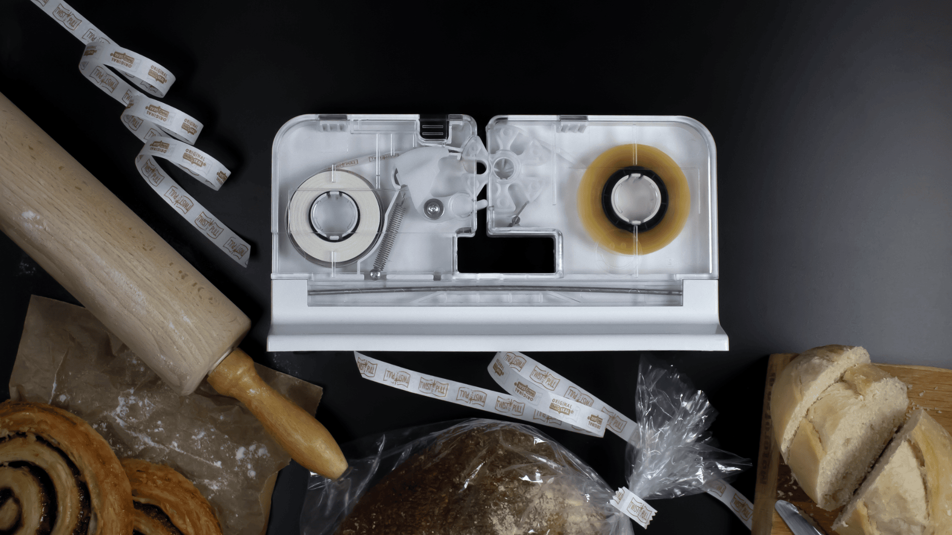 A tabletop labeling machine sits on a black surface surrounded by a rolling pin, pastries, sliced bread, and bread in a clear plastic bag. Two label rolls feed into the machine, with printed labels scattered beside it.