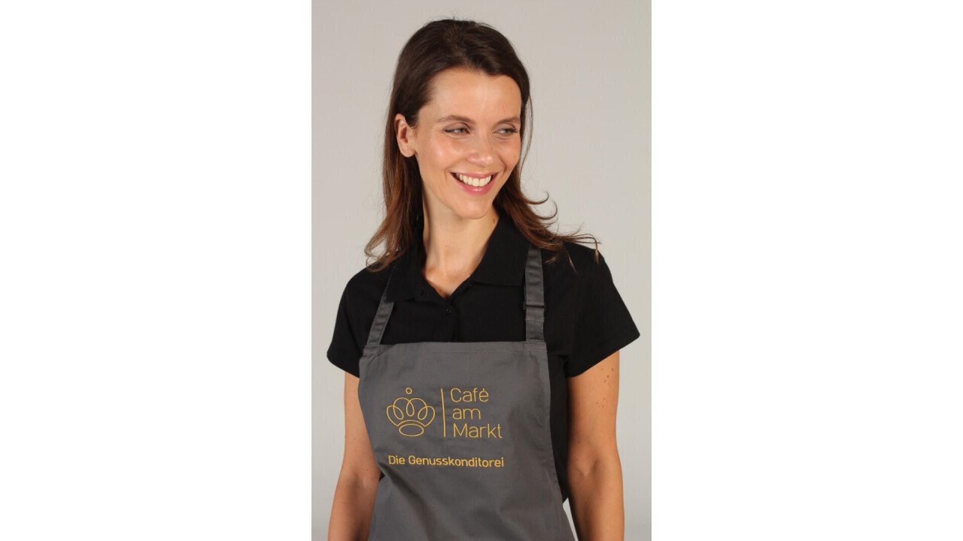 A woman with long brown hair, wearing a black polo shirt and a gray apron that reads Café am Markt Die Genusskonditorei in gold, smiles while looking to her left against a plain background.