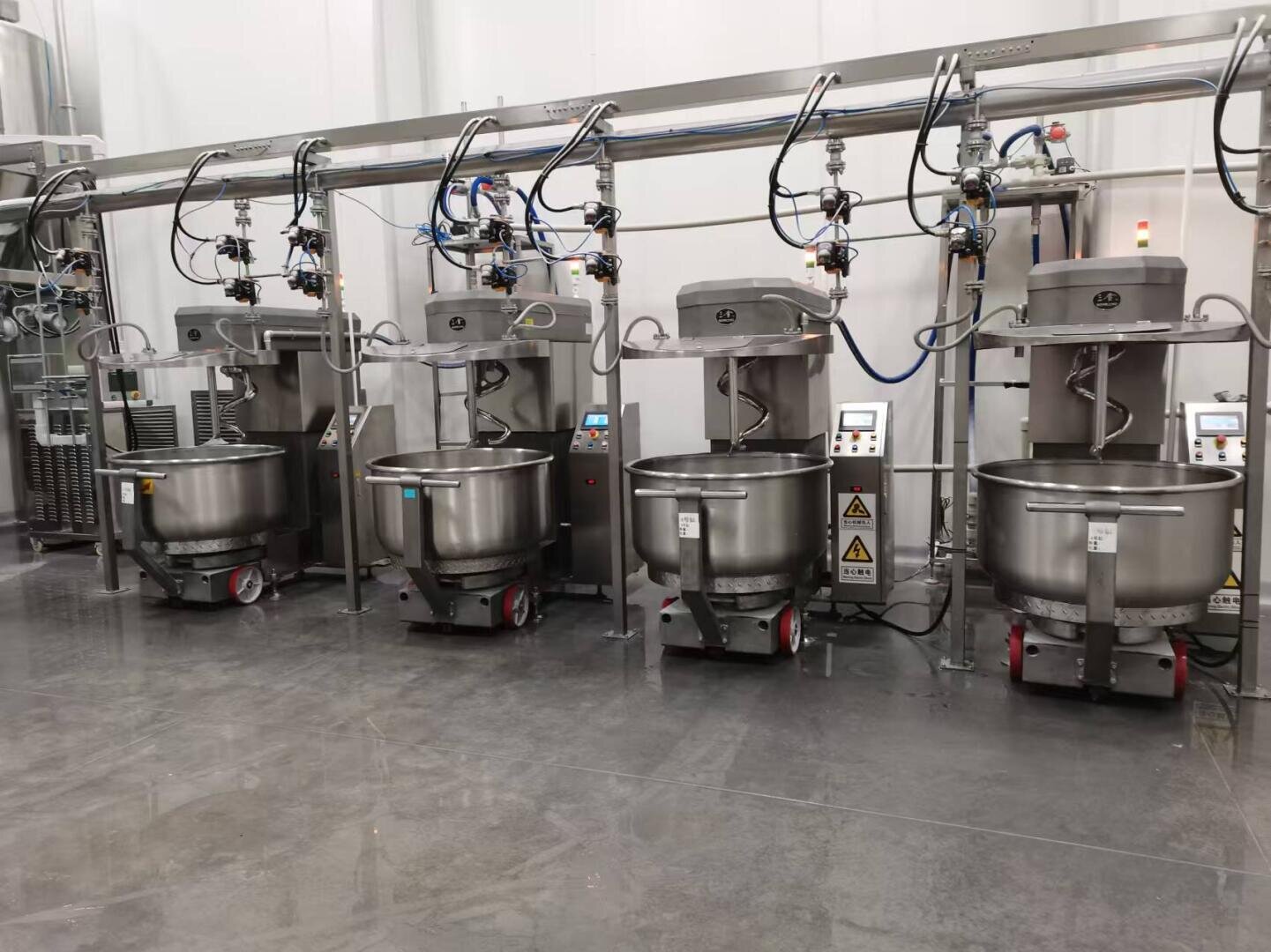 Four large industrial dough mixers with control panels and warning signs are lined up in a clean, modern food processing facility with reflective floors and stainless steel equipment.