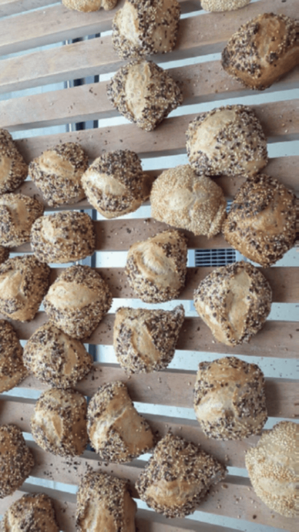 Rows of assorted seeded bread rolls cooling on a wooden rack, with some topped with sesame seeds and others with mixed seeds.