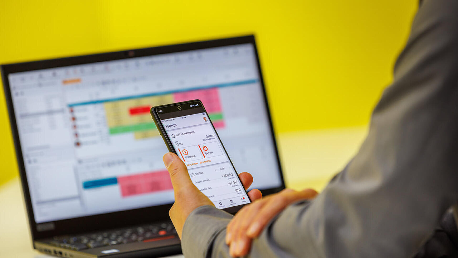 Eine Person in einem grauen Hemd hält ein Smartphone in der Hand, auf dem eine App angezeigt wird, und sitzt vor einem aufgeklappten Laptop, auf dessen Bildschirm eine bunte Tabelle oder ein Zeitplan zu sehen ist, vor einem leuchtend gelben Hintergrund.