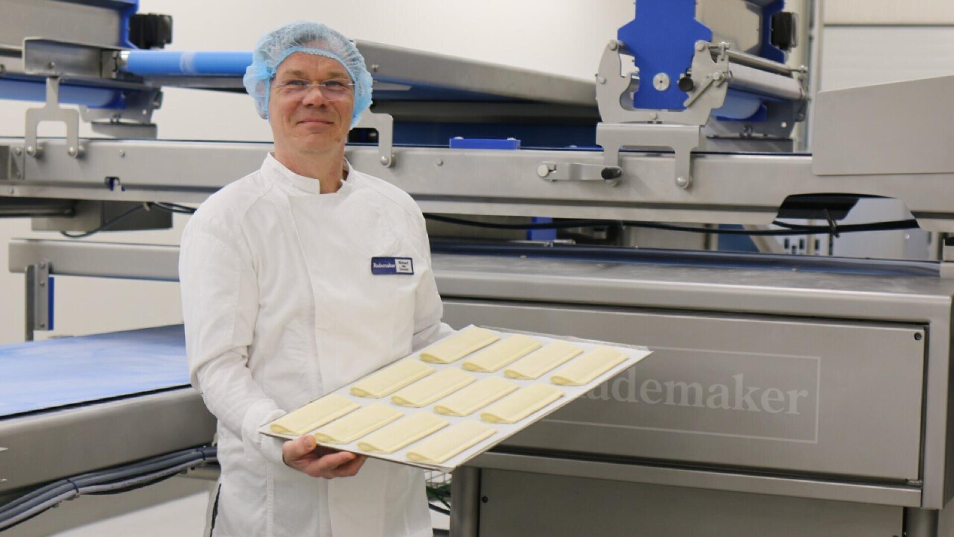 A baker wearing a white coat and blue hairnet holds a tray with neatly arranged unbaked dough strips in front of industrial bakery equipment.