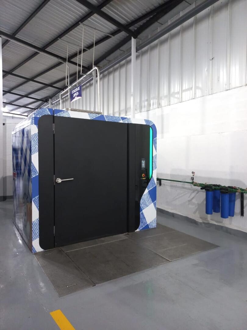 Large, modern industrial booth with a dark door and blue checkered edges inside a spacious, well-lit facility. Nearby, three blue water filters are mounted on the wall with pipes attached.