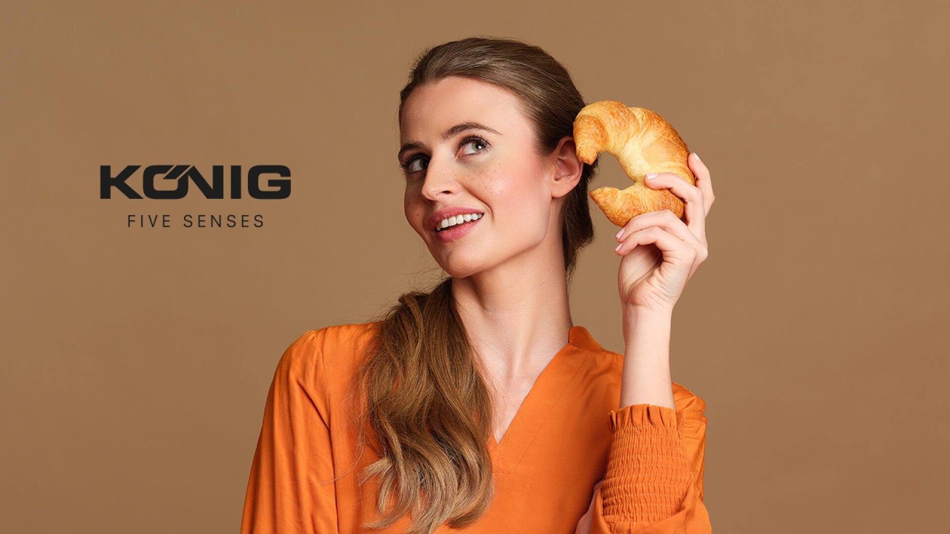 Eine lächelnde Frau in einem orangefarbenen Oberteil hält ein Croissant an ihr Ohr, als ob sie ihm zuhören würde. Der Hintergrund ist beige, und links erscheint die Aufschrift König Five Senses.