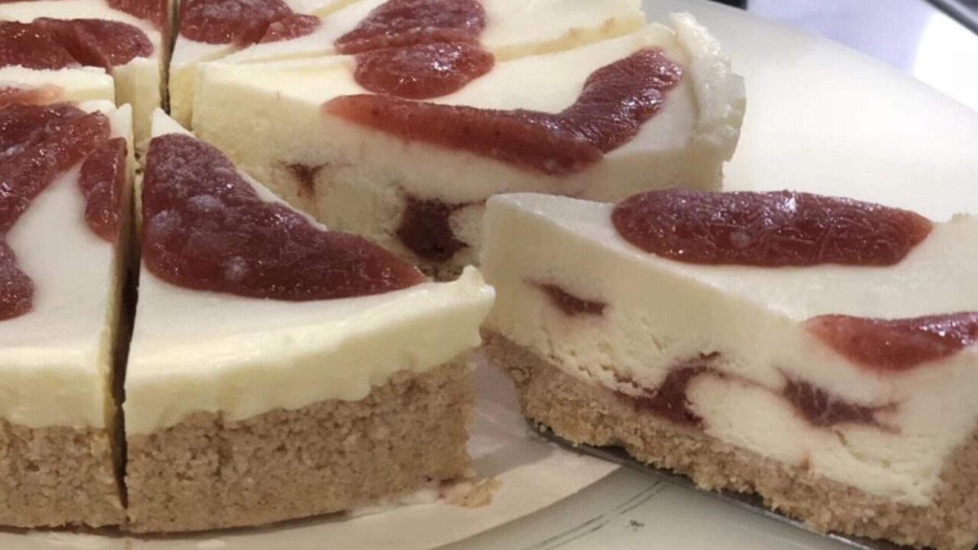 Nahaufnahme eines Käsekuchens mit krümeligem Biskuitboden und roter Fruchtsoße, portioniert auf einem weißen Teller.