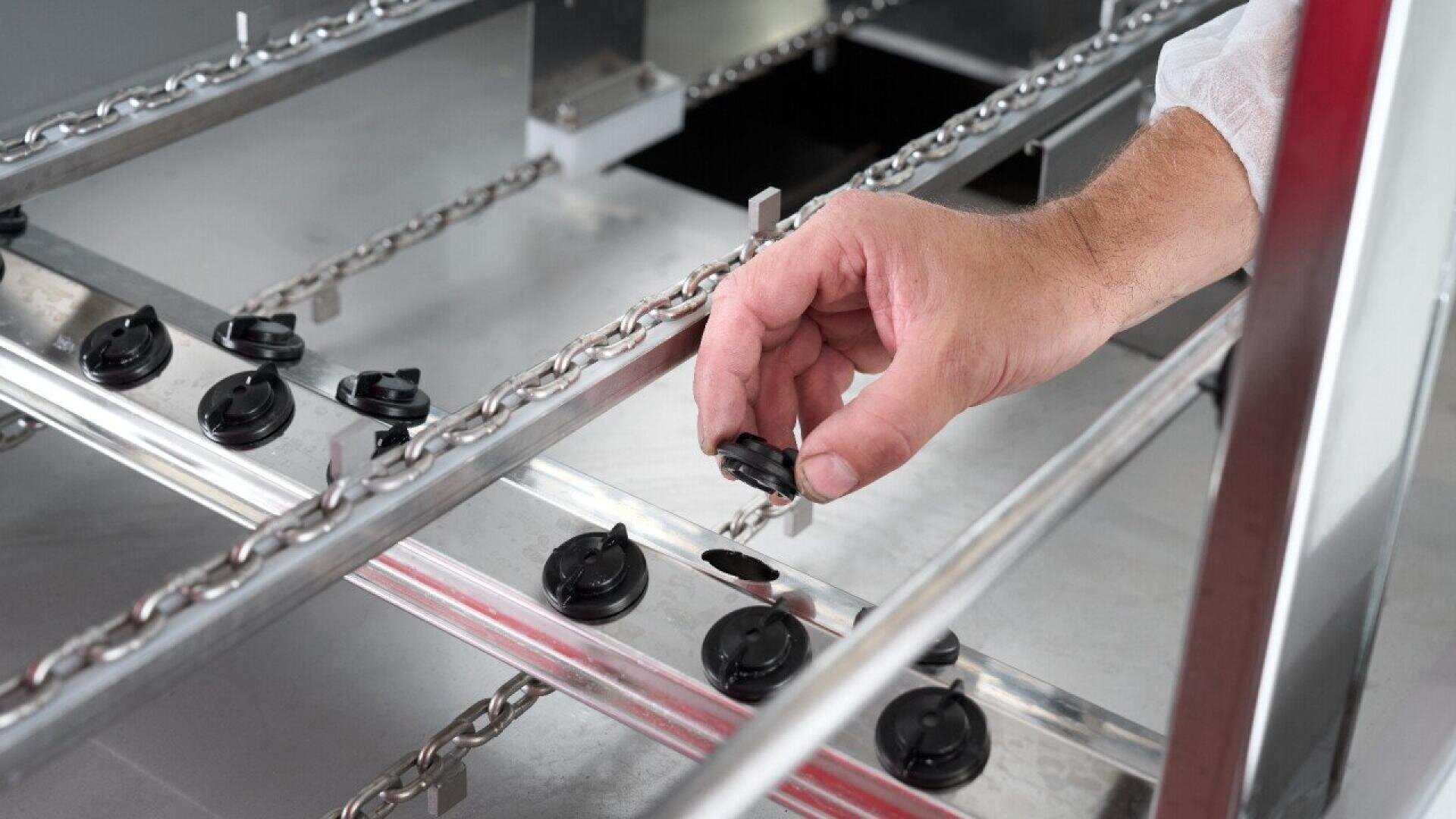 A person’s hand places a black plastic component onto a metal assembly line with chains and rails, in an industrial or manufacturing setting.