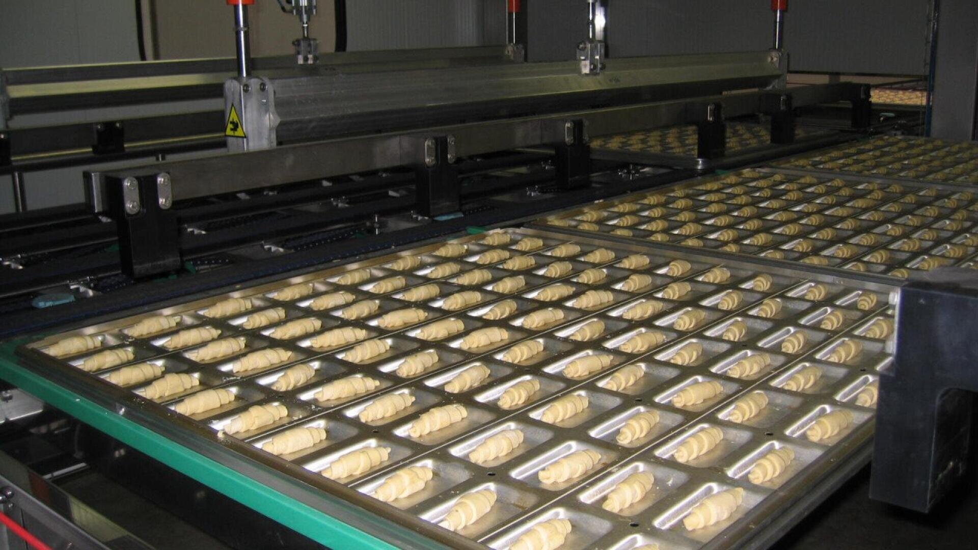 Reihen von ungebackenem, halbmondförmigem Gebäck sind auf großen Metalltabletts angeordnet, die durch eine industrielle Backmaschine in einer Fabrik laufen.