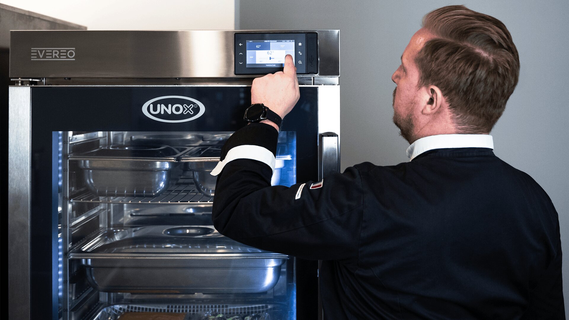 Ein Mann in einer schwarzen Kochjacke bedient das Touchscreen-Bedienfeld eines gewerblichen Küchengeräts aus Edelstahl mit der Aufschrift "UNOX", in dessen Inneren Metallschalen zu sehen sind.