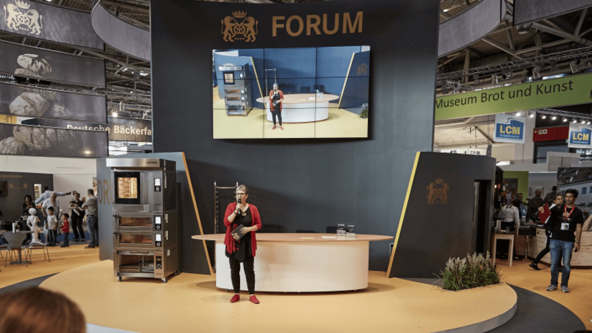 Eine Frau in Rot und Schwarz steht auf einer Bühne mit einer leeren weißen Theke vor ihr. Hinter ihr befindet sich ein großer Bildschirm, der ihr Bild zeigt, und ein Schild mit der Aufschrift "FORUM". Menschen besuchen die umliegenden Stände.