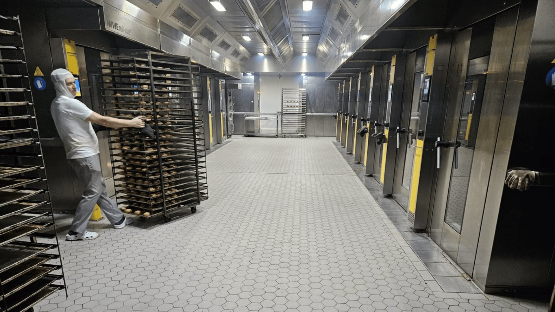 Eine Person in Schutzkleidung schiebt ein hohes Regal mit Backblechen in eine große, moderne Industriebäckerei, in der mehrere Öfen auf beiden Seiten des geräumigen, gekachelten Raums stehen.