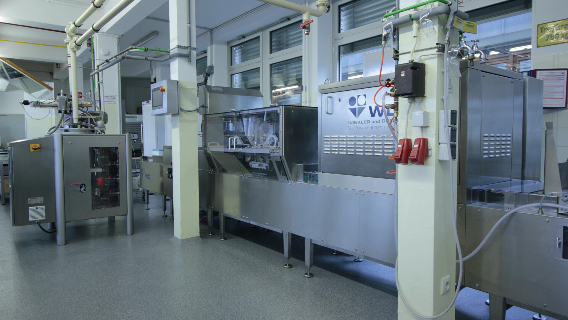 Eine moderne, industrielle Lebensmittelverpackungsmaschine in einer sauberen, gut beleuchteten Anlage mit Edelstahlausrüstung, Bedienfeldern und sichtbaren Sicherheitsfunktionen. Große Fenster sorgen für Tageslicht.