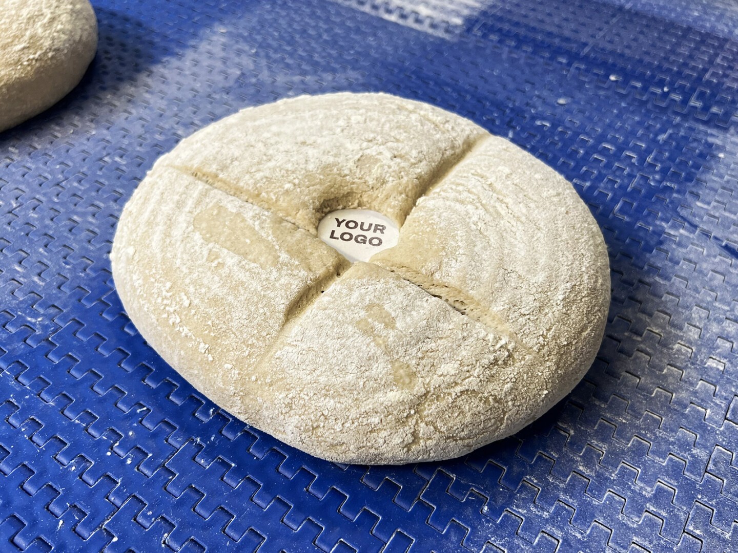 A round, uncooked loaf of bread with a cross-shaped cut on top sits on a blue textured surface. A small circular label at the center of the loaf reads YOUR LOGO.