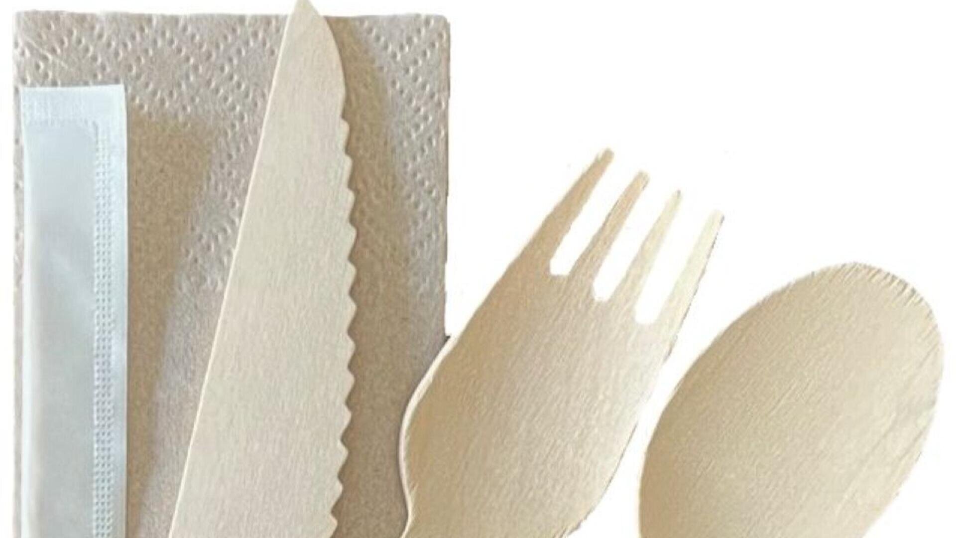 Ein Satz umweltfreundlicher Einwegutensilien, einschließlich Messer, Gabel und Löffel, wird neben einer Serviette und einem kleinen Päckchen, das möglicherweise einen Zahnstocher oder ein Feuchttuch enthält, auf einem weißen Hintergrund platziert.
