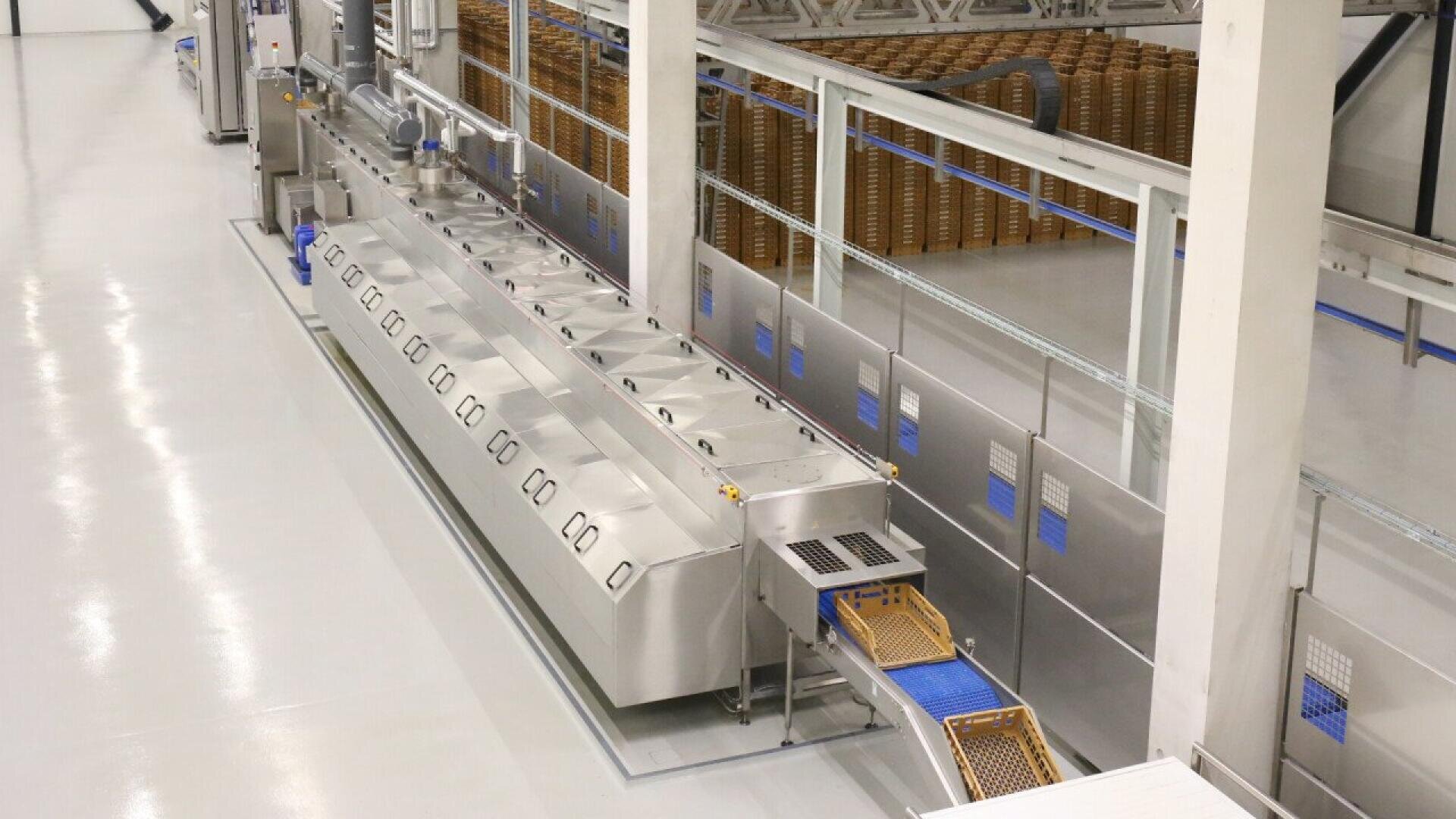 A modern industrial food processing line with stainless steel machines and conveyor belts, transporting trays of food products in a clean, brightly lit factory environment.