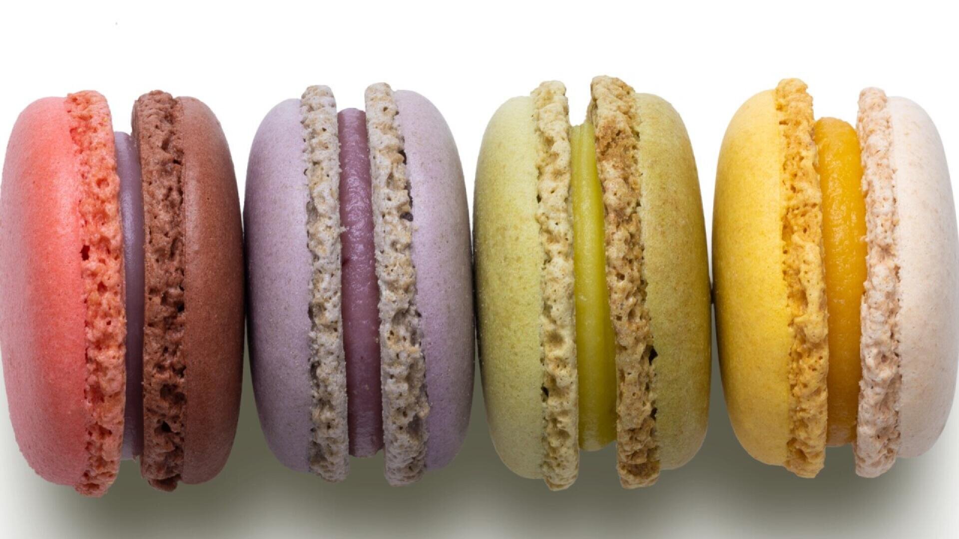 Five colorful macarons, arranged in a row on a white background. The macarons are red, purple, green, yellow, and light beige, each with a creamy filling visible between their two cookie-like halves.