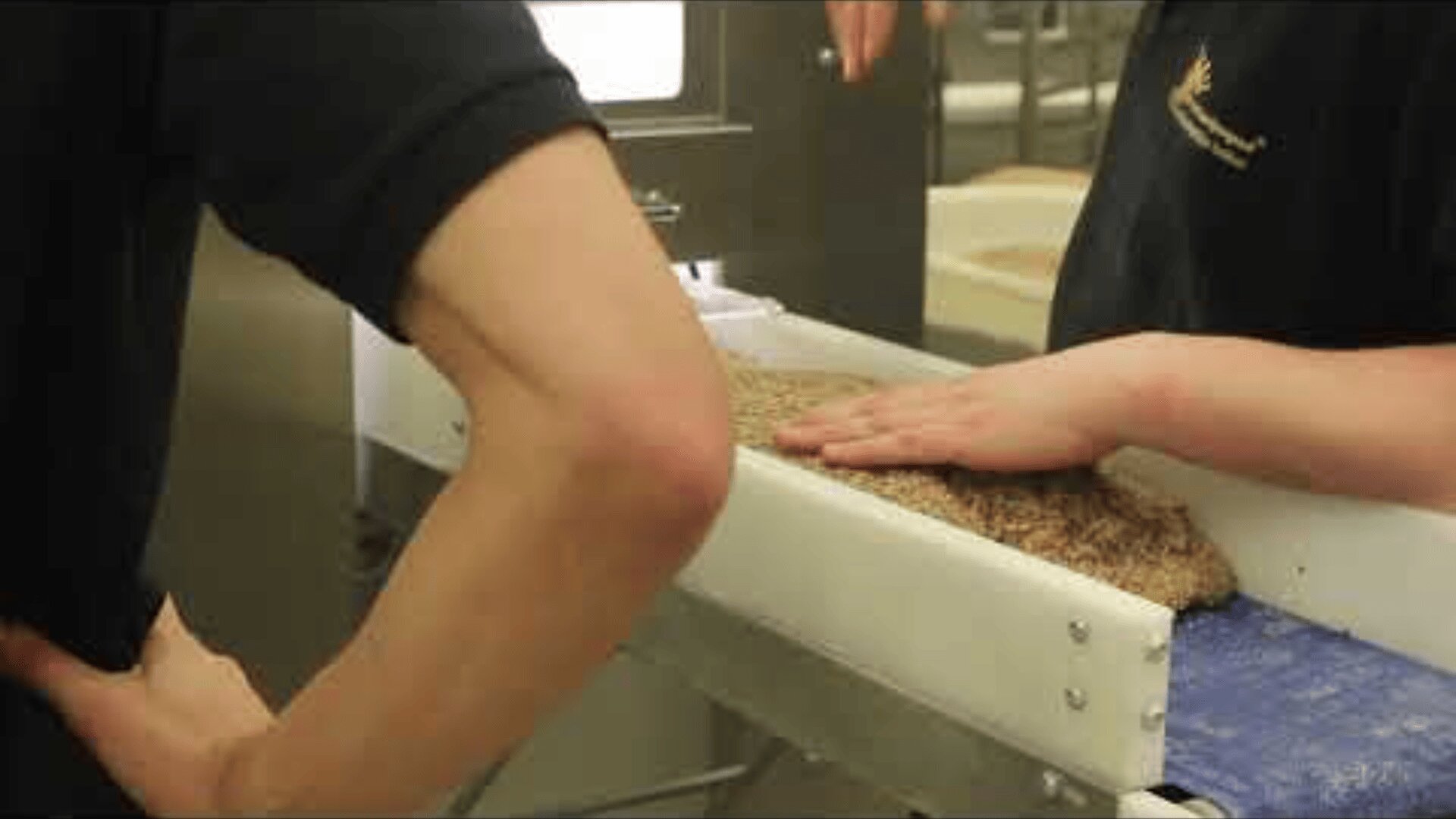 Zwei Personen arbeiten an einem Förderband; eine Person drückt mit einer Hand auf ein flaches, strukturiertes braunes Material auf dem Band, während die andere mit einem in der Hüfte angewinkelten Arm daneben steht.
