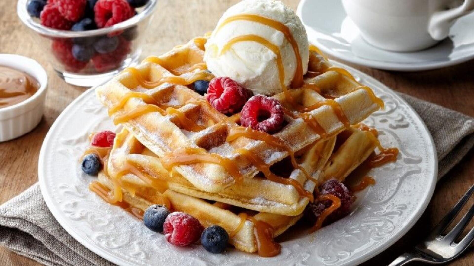 Ein Teller mit Waffeln, die mit Vanilleeis, frischen Himbeeren und Blaubeeren belegt und mit Karamellsauce beträufelt sind. Im Hintergrund stehen eine Tasse, eine Schale mit Beeren und eine Schale mit Karamell.