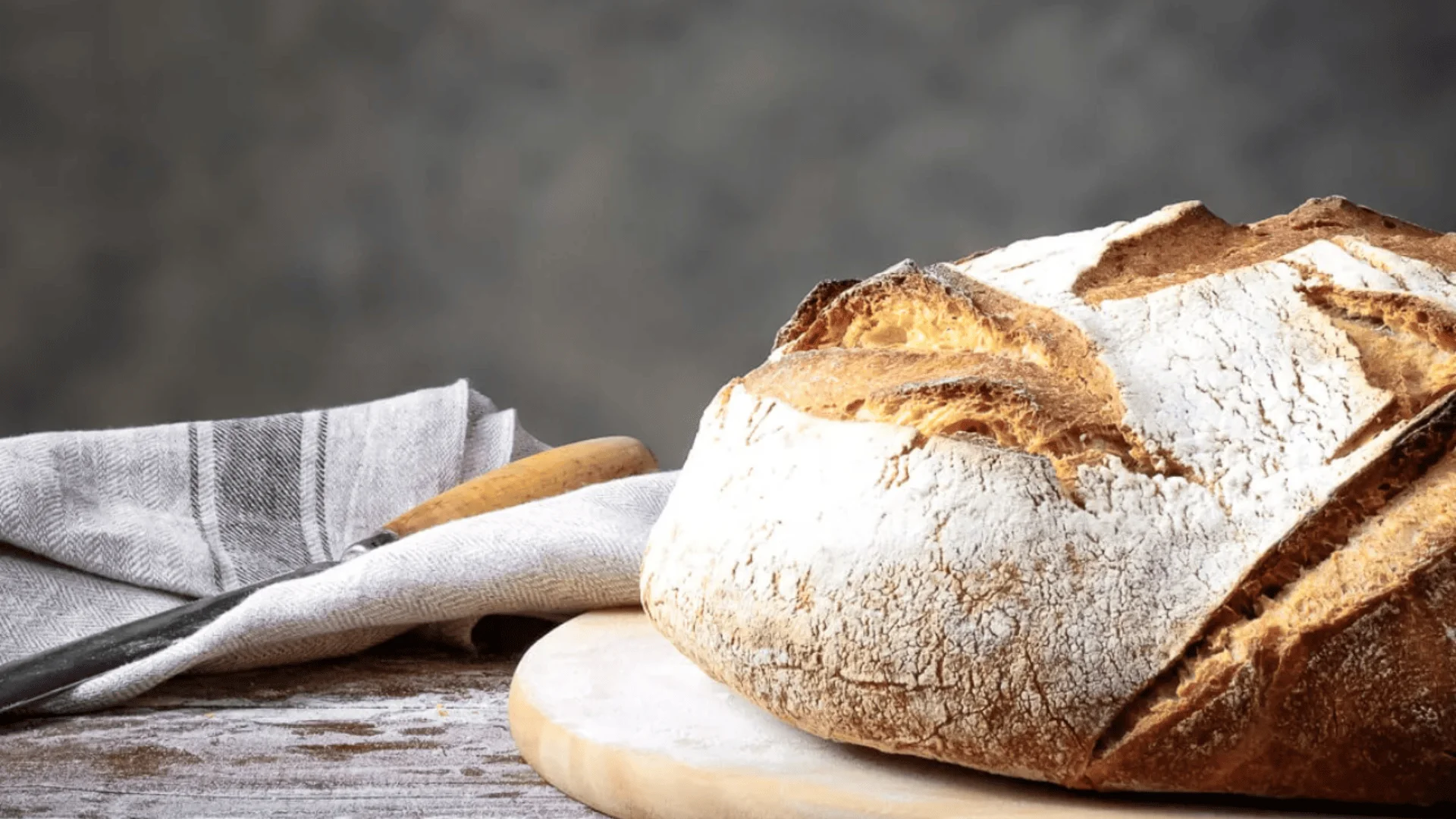 Ein rustikaler, runder Laib knusprigen Handwerksbrotes liegt auf einem hölzernen Schneidebrett, neben einem gestreiften Tuch und einem Brotmesser, vor einem neutral strukturierten Hintergrund.