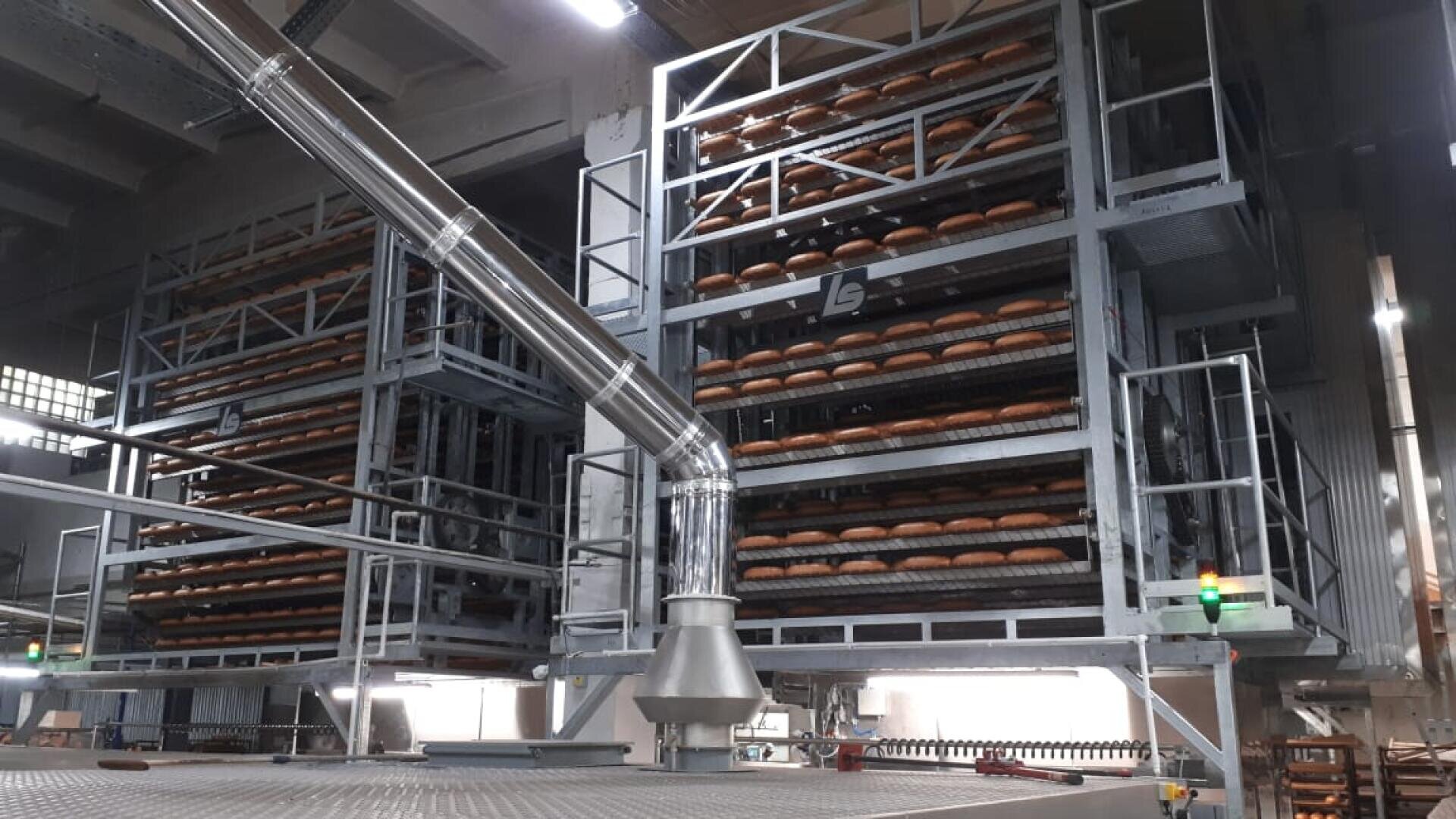 Industrial bakery scene showing large metal racks filled with rows of baked bread loaves. Metal pipes and machinery are visible, indicating an automated bread production process inside a factory.