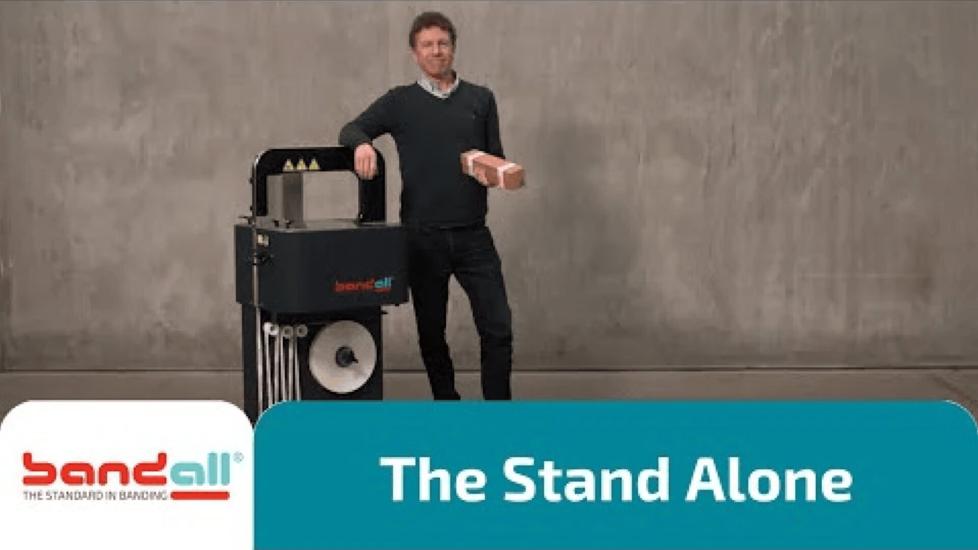 Ein Mann steht neben einer Bandall-Umreifungsmaschine und hält einen ziegelsteinförmigen Gegenstand. Der Hintergrund ist einfarbig grau, und der Text auf dem Bild lautet: "The Stand Alone". Das Bandall-Logo erscheint in der unteren linken Ecke.
