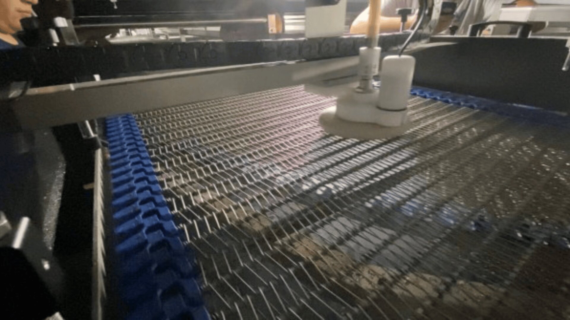 Nahaufnahme einer Maschine mit Metalldrähten und blauen Förderbandsegmenten, wahrscheinlich Teil einer industriellen Lebensmittelverarbeitungs- oder Fertigungsstraße. Weiße Komponenten und oben liegende Maschinen sind sichtbar.