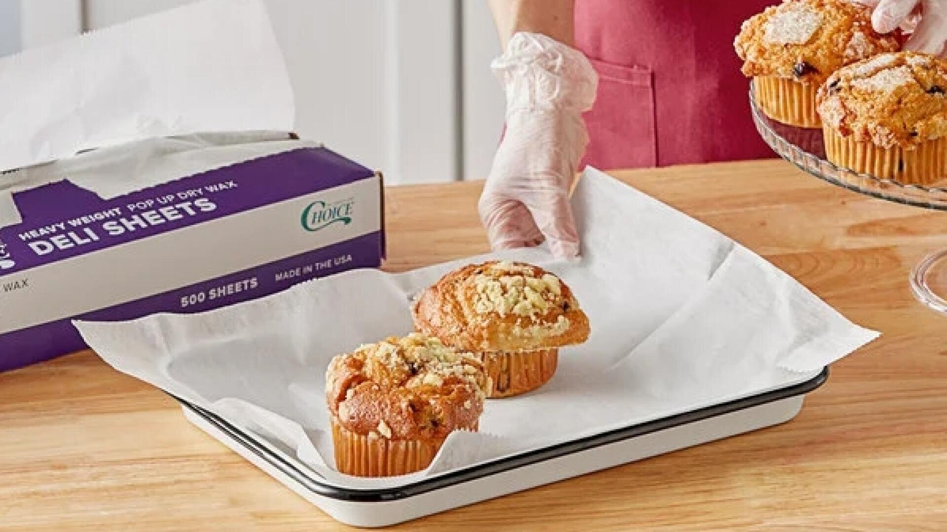 A person wearing gloves places muffins onto a parchment-lined tray on a wooden table. More muffins are arranged on a glass cake stand nearby, and a box of deli sheets is in the background.