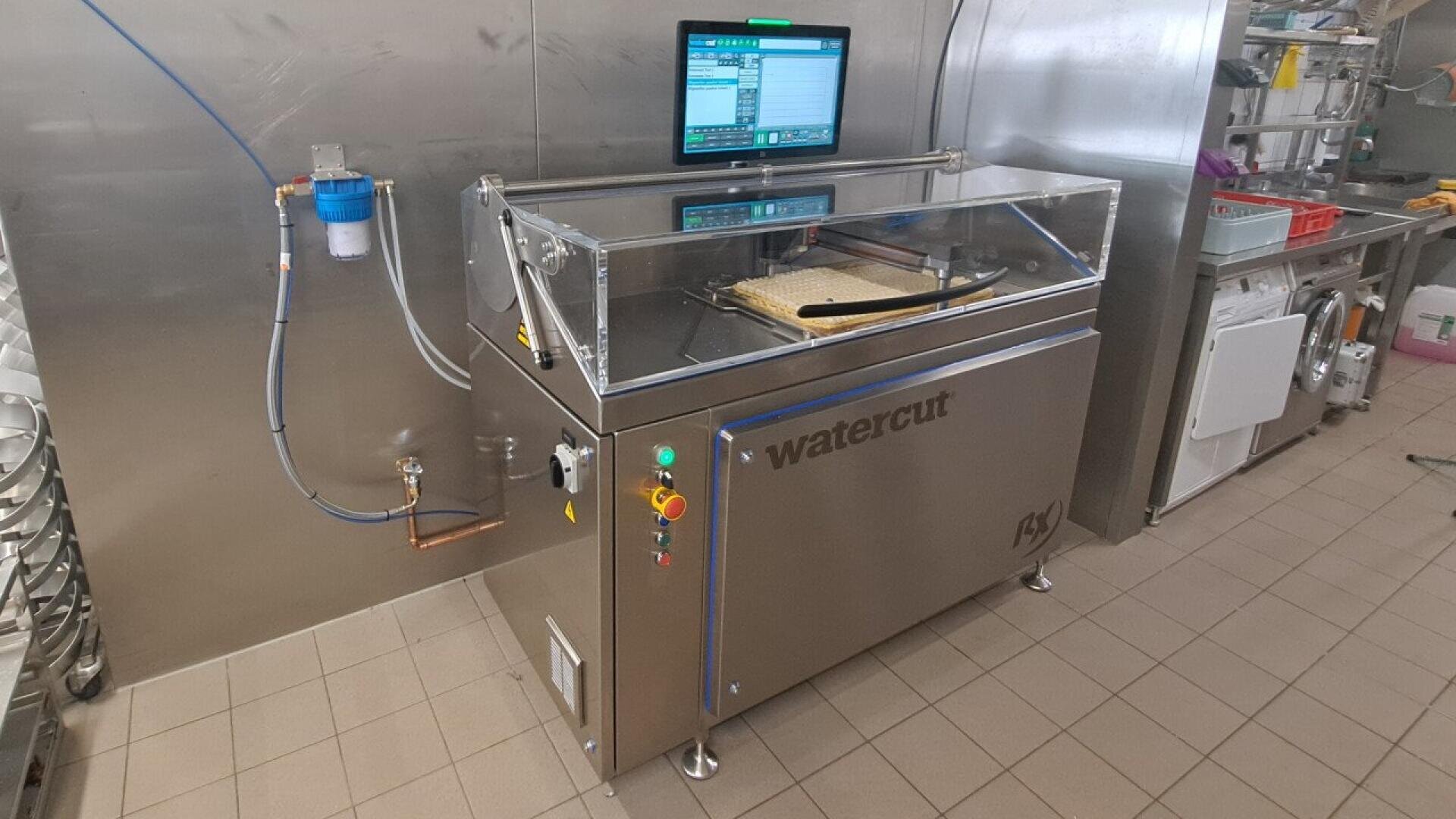 Eine Watercut-Maschine aus Edelstahl mit einer transparenten Abdeckung und einem Bedienfeld, die zum präzisen Schneiden von Lebensmitteln verwendet wird, steht in einer Großküche mit gefliesten Böden und Wänden. Ein Computermonitor zeigt die Bedienelemente über der Maschine an.